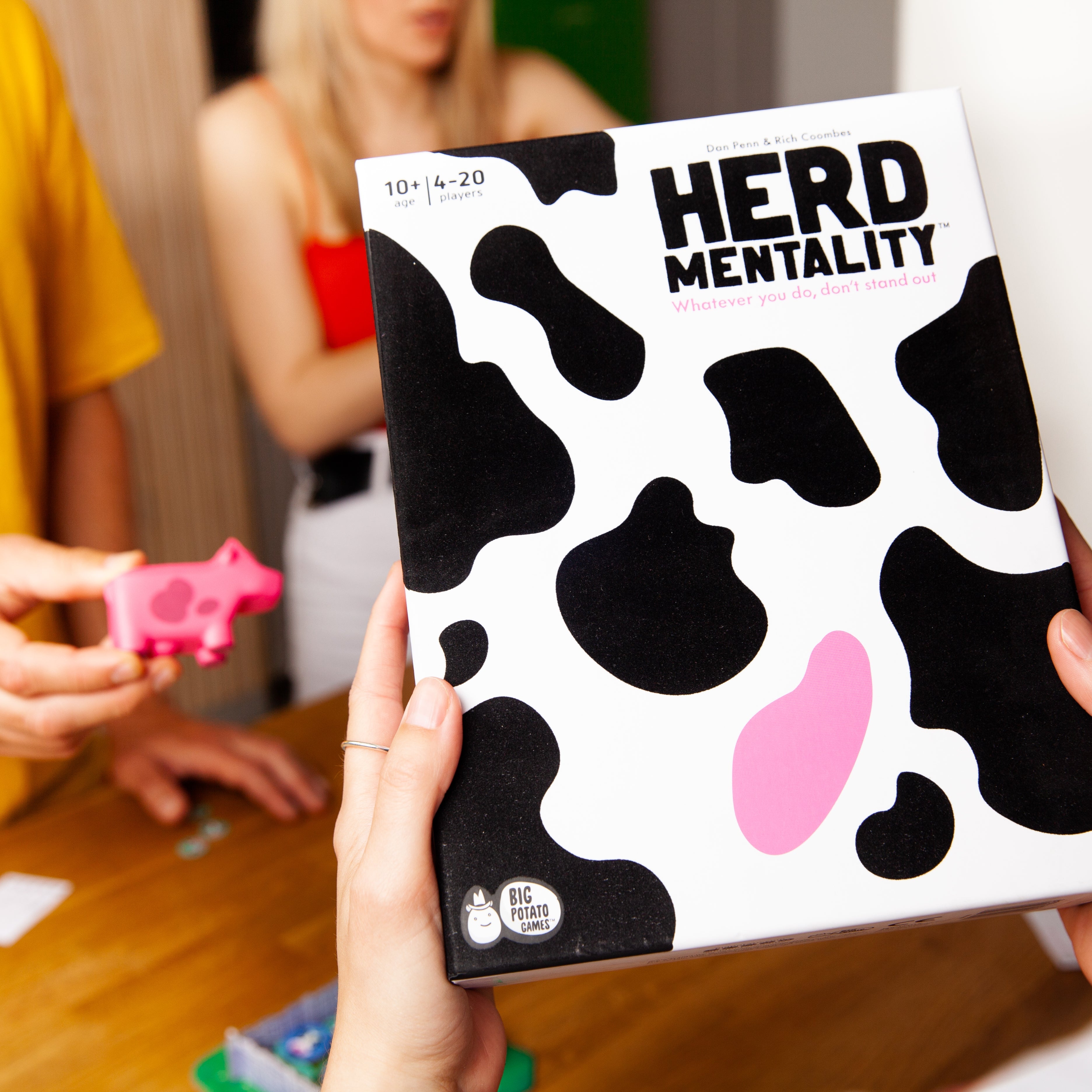 Board game box titled 'HERD MENTALITY' held by a person with people in the background.